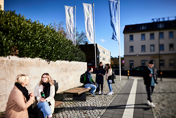 Studierende sitzen und stehen vor dem Eingang der Hochschule Fulda, Fahnen der Hochschule wehen im Wind